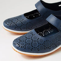 A pair of navy blue Mary Jane-style shoes with floral laser-cut perforations on the toe and upper straps, featuring a white midsole and a natural brown outsole, shown on a light gray background.