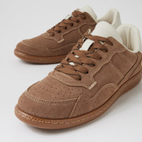 A pair of low-top brown suede sneakers with matching brown laces, white interior lining, and a gum-colored sole, photographed on a clean light gray studio background.