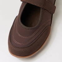 A close-up of a dark brown slip-on shoe with a hook-and-loop strap across the top, featuring decorative contrast stitching and a gum outsole, shown on a light gray background.